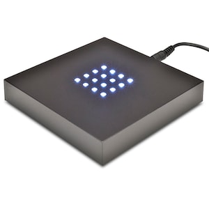 May include: A black, square LED display base with a grid of bright white lights. The base has a matte finish and a power cord extending from the side. The lights are arranged in a diamond pattern.