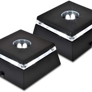May include: Two black rotating display bases with white LED lights. The bases are square and have a circular platform in the center.