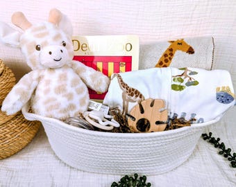 May include: A white rope basket filled with baby items. Includes a plush giraffe, a Dear Zoo book, a giraffe-themed blanket, a wooden teether, and a baby bib with giraffe and tree illustrations. The basket is on a white surface.