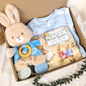 May include: A gift box containing a plush bunny, a blue baby onesie, and a copy of "The Tale of Peter Rabbit" storybook. The bunny has a blue outfit and a yellow ring. The onesie has a pattern of white shapes.