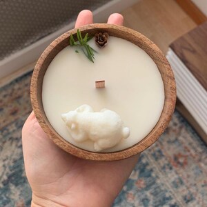 May include: A white candle in a wooden bowl with a small white bear figurine on top. The candle is surrounded by green sprigs and a small brown pinecone.