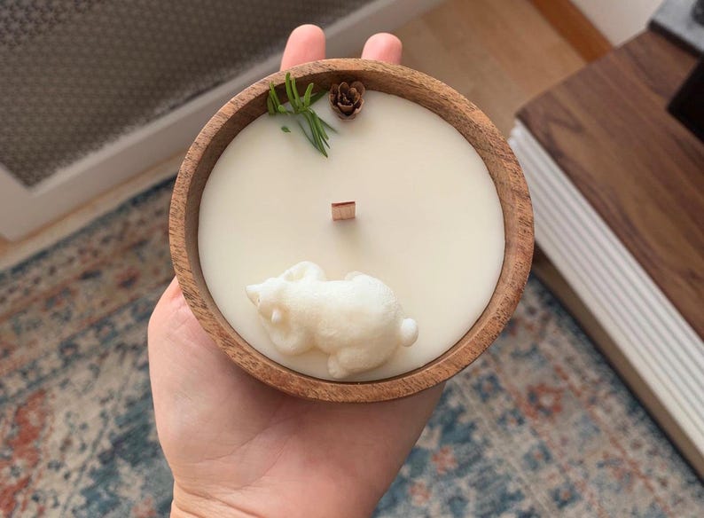 May include: A white candle in a wooden bowl with a small white bear figurine on top. The candle is surrounded by green sprigs and a small brown pinecone.
