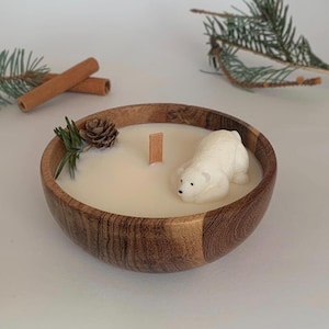 May include: A white polar bear candle in a wooden bowl with a white wax base. The candle is surrounded by pine needles and a small pine cone.