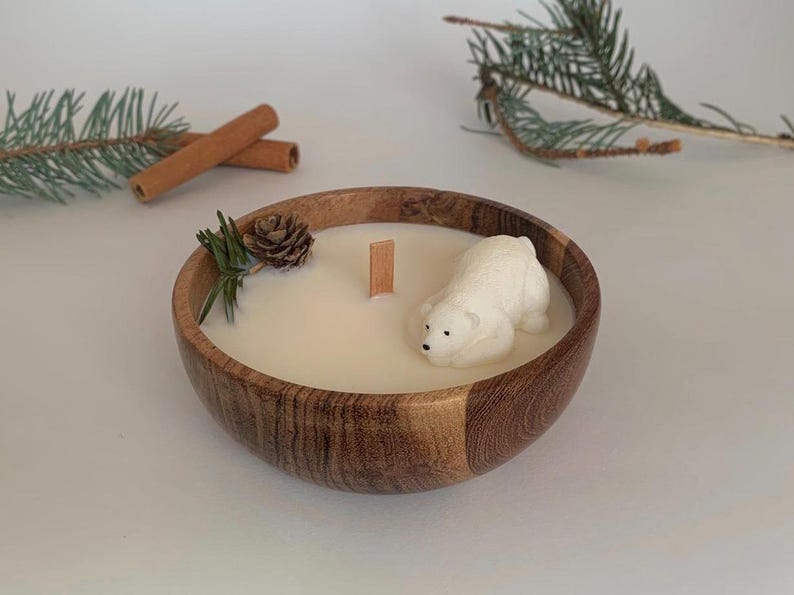 May include: A white polar bear candle in a wooden bowl with a white wax base. The candle is surrounded by pine needles and a small pine cone.