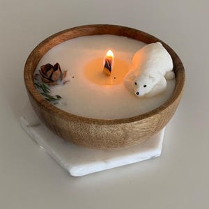 May include: A white candle in a wooden bowl with a polar bear figurine and a pine cone. The candle is lit and the wax is melted.