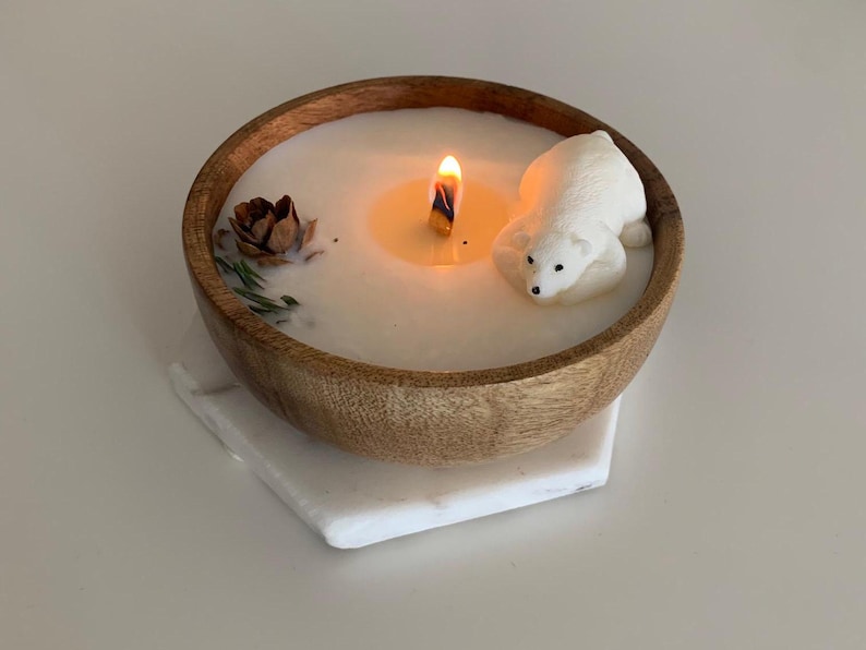 May include: A white candle in a wooden bowl with a polar bear figurine and a pine cone. The candle is lit and the wax is melted.