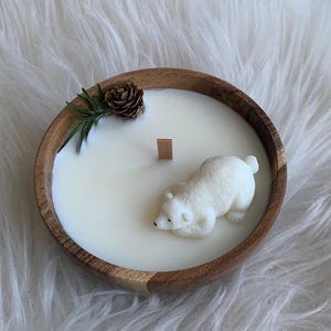 May include: A white candle in a wooden bowl with a polar bear figurine on top. The candle is surrounded by a sprig of greenery and a pine cone.