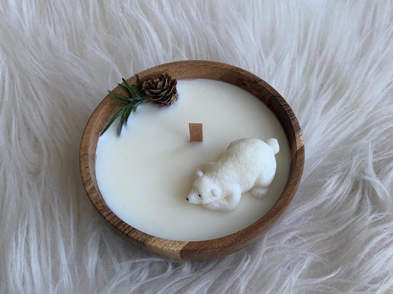May include: A white candle in a wooden bowl with a polar bear figurine on top. The candle is surrounded by a sprig of greenery and a pine cone.