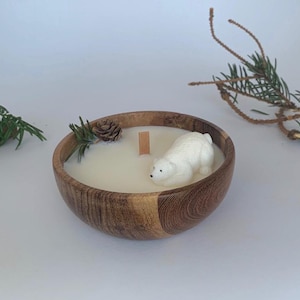 May include: A white polar bear candle in a wooden bowl with a white wax pool and a wooden wick. The bowl is surrounded by pine needles and a pine cone.