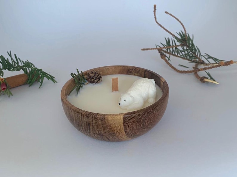 May include: A white polar bear candle in a wooden bowl with a white wax pool and a wooden wick. The bowl is surrounded by pine needles and a pine cone.