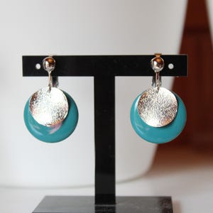 May include: A pair of teal and silver earrings on a black stand. Each earring has a textured silver disc over a teal circle. The earrings are clip-on style. The earrings are displayed on a black stand.