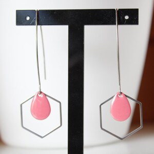 May include: A pair of drop earrings with a geometric design. Each earring has a silver-tone, open hexagon shape with a pink teardrop-shaped accent in the centre. The earrings hang from a thin silver wire.