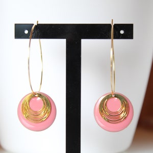 May include: A pair of gold hoop earrings featuring a dangling pink circular disc. Each disc is accented with a smaller, layered gold circular charm that has a series of concentric rings. The earrings are displayed on a black T-shaped stand.
