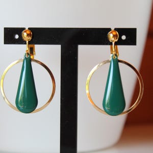 Peut inclure: Boucles d'oreilles à clip avec des cercles dorés et des pendentifs en forme de goutte d'eau vert foncé. Les boucles d'oreilles sont présentées sur un support noir. Finition polie. Accessoire de mode élégant.