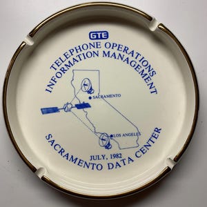 May include: A white ceramic ashtray with a gold rim. The ashtray features blue text and a map of California with the words "GTE TELEPHONE OPERATIONS INFORMATION MANAGEMENT SACRAMENTO DATA CENTER JULY, 1982."