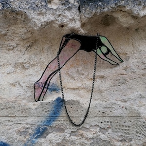 Może przedstawiać: Witraż wiszący na czarnym łańcuchu. Witraż przedstawia osobę nurkującą głową naprzód do wody. Osoba jest różowa i zielona.