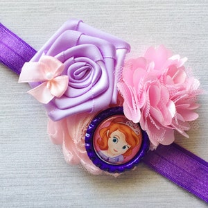 May include: Purple elastic headband with a lavender satin flower, a pink tulle flower, and a purple button with a picture of a princess.