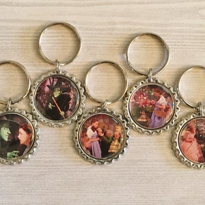 May include: Set of five silver keychains with round, silver-colored bottle cap charms. Each charm features a different image from the Wizard of Oz movie. The images include Dorothy, the Wicked Witch of the West, and the poppy field.