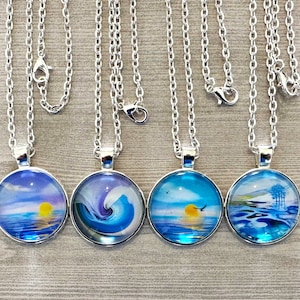 May include: Five silver necklaces with round glass pendants featuring different ocean-themed designs. The pendants depict a sunset over the ocean, a crashing wave, a sunrise over the ocean, a bird flying over the ocean, and a mountain range reflected in the water.