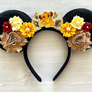 May include: Black mouse ears with a brown, yellow, and red flower embellishment. A cartoon lion character is attached to the headband.