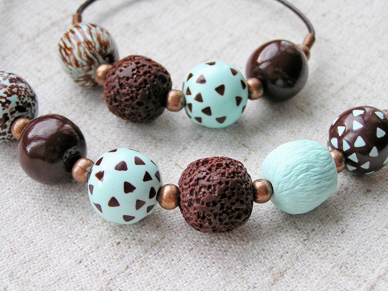 Op de afbeelding: Een bruine en turquoise kralenketting met bruine en witte gepatroneerde kralen. De ketting is gemaakt van verschillende materialen, waaronder hout, lavastenen en polymer clay.