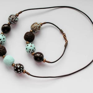 Op de afbeelding: Een bruine leren ketting met een bijpassend armband. De ketting en het armband zijn versierd met bruine en blauwe kralen met patronen.