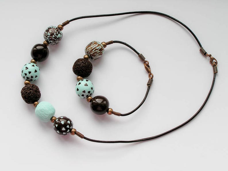 Op de afbeelding: Een bruine leren ketting met een bijpassend armband. De ketting en het armband zijn versierd met bruine en blauwe kralen met patronen.