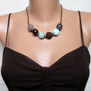 Op de afbeelding: Een bruine en turquoise ketting met ronde kralen. De kralen zijn gemaakt van verschillende materialen, waaronder hout, lavasteen en beschilderd keramiek. De ketting is aan een bruin koord.