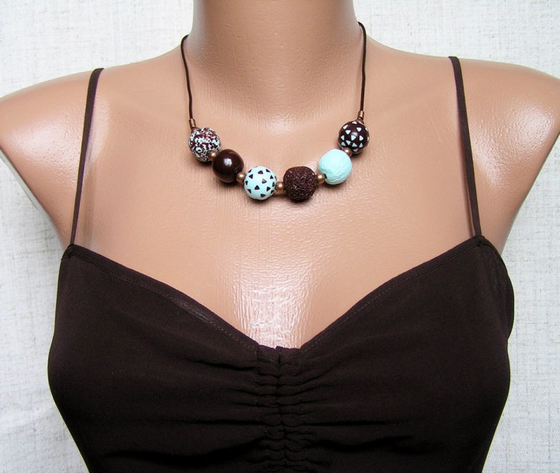 Op de afbeelding: Een bruine en turquoise ketting met ronde kralen. De kralen zijn gemaakt van verschillende materialen, waaronder hout, lavasteen en beschilderd keramiek. De ketting is aan een bruin koord.