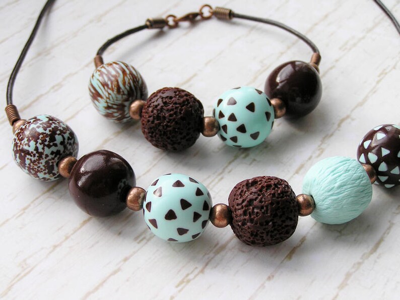 Op de afbeelding: Een bruine en turquoise kralenketting met bruine en witte kralen met patronen. De ketting is gemaakt van verschillende texturen, waaronder gladde, getextureerde en gepatroneerde kralen.