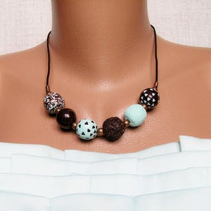 Op de afbeelding: Een bruine en mintgroene kralenketting met een bruin koord. De kralen zijn rond en hebben een geometrisch patroon.