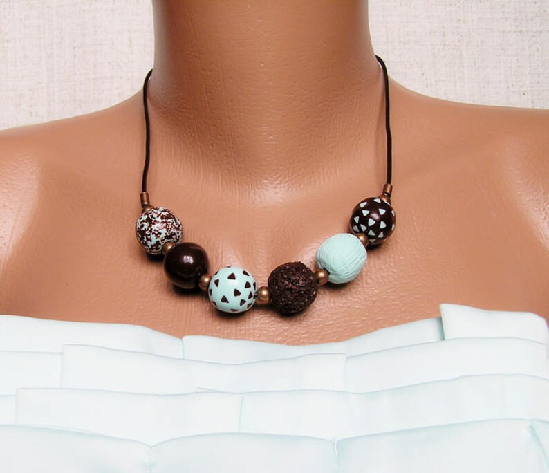 Op de afbeelding: Een bruine en mintgroene kralenketting met een bruin koord. De kralen zijn rond en hebben een geometrisch patroon.