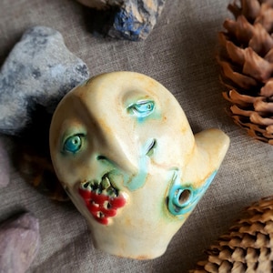 May include: A quirky ceramic face sculpture with turquoise eyes and accents, and a red berry cluster. The sculpture is off-white with blue and green details. Pine cones and rocks are in the background.