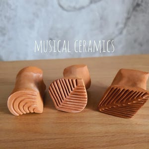 May include: Three terracotta-coloured ceramic stamps with carved patterns on their bases. The left stamp features concentric curved lines, while the centre and right stamps display leaf-like vein textures. The text MUSICAL CERAMICS is visible.