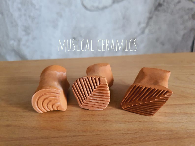 May include: Three terracotta-coloured ceramic stamps with carved patterns on their bases. The left stamp features concentric curved lines, while the centre and right stamps display leaf-like vein textures. The text MUSICAL CERAMICS is visible.