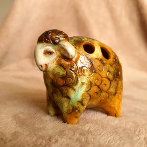 May include: A ceramic sheep ornament with a brown head and a body in shades of yellow, orange, and green. The sheep has two holes on its back. The ornament is on a soft, light brown surface.