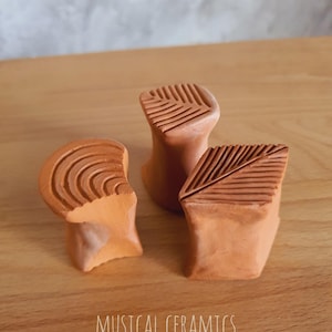 May include: Three terracotta-coloured ceramic objects with textured tops, including one with concentric circles and two with linear patterns. The text MUSICAL CERAMICS is printed at the bottom of the image. These small, hand-formed clay pieces feature distinct geometric carvings on their upper surfaces.