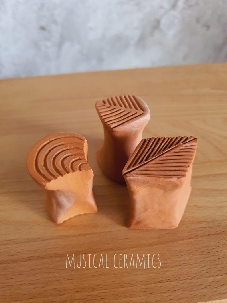 May include: Three terracotta-coloured ceramic objects with textured tops, including one with concentric circles and two with linear patterns. The text MUSICAL CERAMICS is printed at the bottom of the image. These small, hand-formed clay pieces feature distinct geometric carvings on their upper surfaces.