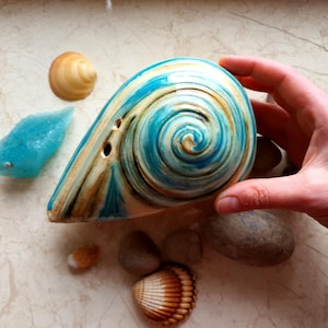 May include: A ceramic ocarina in a spiral seashell design, with blue, white, and brown colours. The ocarina is surrounded by small seashells, pebbles, and a blue crystal-like object. The ocarina is held by a hand.
