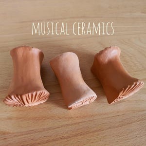 May include: Three terracotta-coloured ceramic objects with flared ends and textured bases, arranged in a row. The text MUSICAL CERAMICS is printed in white above the objects. Each piece has a smooth, cylindrical body that widens at both ends.