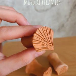 May include: Terracotta ceramic stamps with a fan-shaped ginkgo leaf pattern on the face. The stamps have a cylindrical handle and a textured, carved surface. The text MUSICAL CERAMICS is visible at the top of the image.