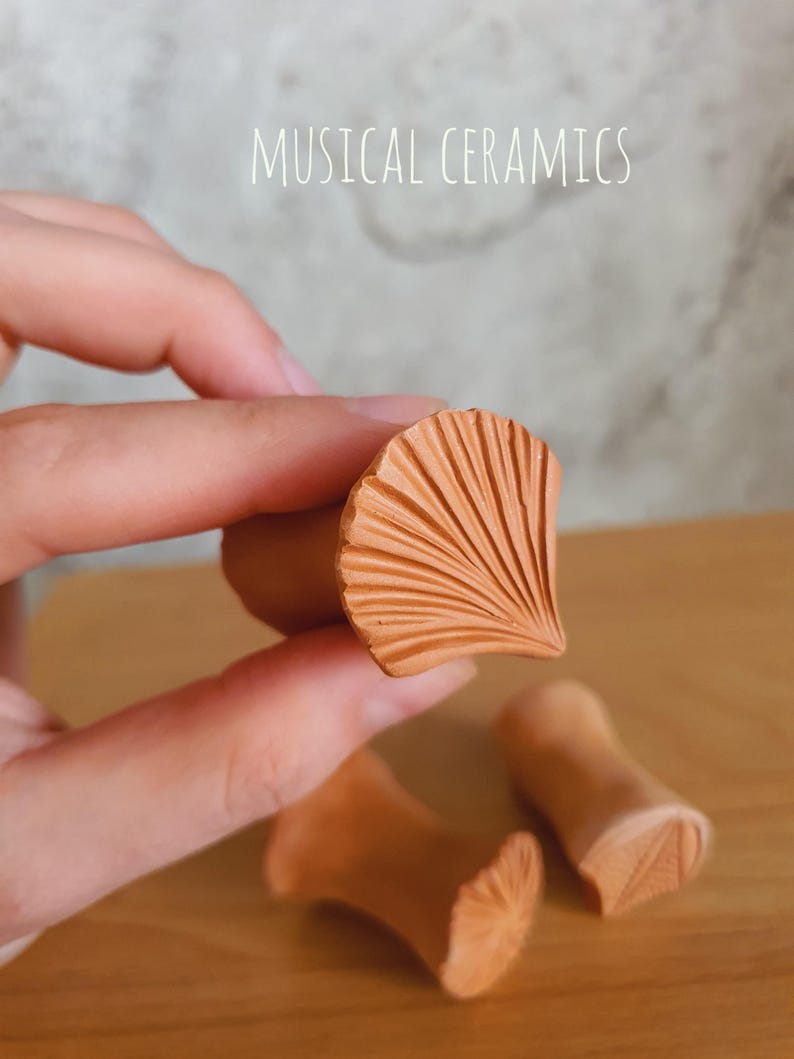 May include: Terracotta ceramic stamps with a fan-shaped ginkgo leaf pattern on the face. The stamps have a cylindrical handle and a textured, carved surface. The text MUSICAL CERAMICS is visible at the top of the image.