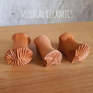 May include: Three terracotta clay stamps with the text MUSICAL CERAMICS visible above. Each stamp features a unique carved pattern on the base, including a radial fan design, a four-leaf floral motif, and a textured shell-like pattern.