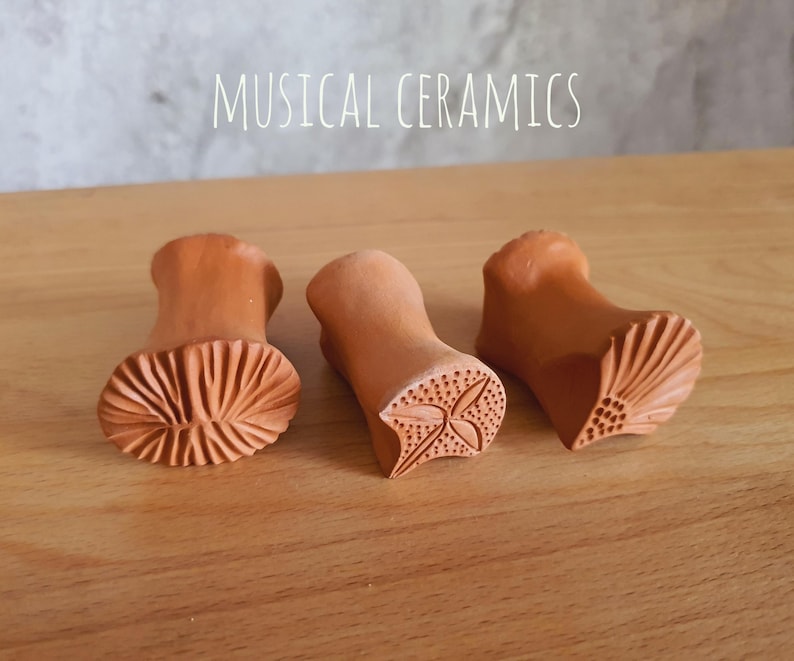 May include: Three terracotta clay stamps with the text MUSICAL CERAMICS visible above. Each stamp features a unique carved pattern on the base, including a radial fan design, a four-leaf floral motif, and a textured shell-like pattern.