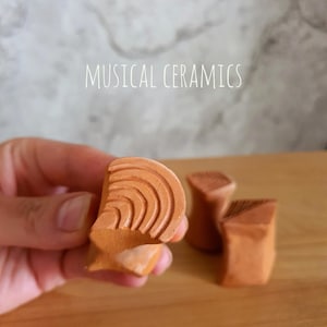 May include: Three small terracotta-coloured ceramic objects with textured surfaces. One object is held in the foreground, displaying concentric curved lines, while two others sit behind it. The text MUSICAL CERAMICS is visible above.