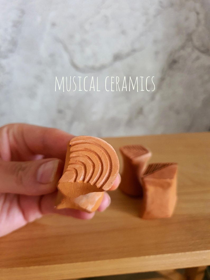May include: Three small terracotta-coloured ceramic objects with textured surfaces. One object is held in the foreground, displaying concentric curved lines, while two others sit behind it. The text MUSICAL CERAMICS is visible above.