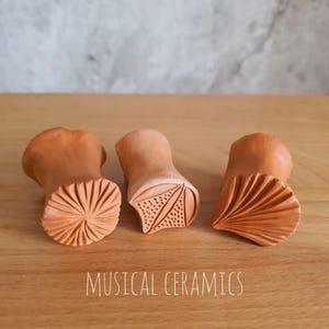 May include: Three terracotta clay stamps with distinct carved patterns on their faces, including a radial sunburst, a triangular leaf-like design with dots, and a fan-shaped motif. The text MUSICAL CERAMICS is printed at the bottom.