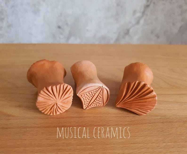 May include: Three terracotta clay stamps with distinct carved patterns on their faces, including a radial sunburst, a triangular leaf-like design with dots, and a fan-shaped motif. The text MUSICAL CERAMICS is printed at the bottom.