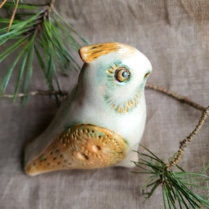 May include: A ceramic bird figurine in light blue and cream, with brown and gold accents. The bird is near pine needles and a pine cone. The wing has a textured design. A decorative item.