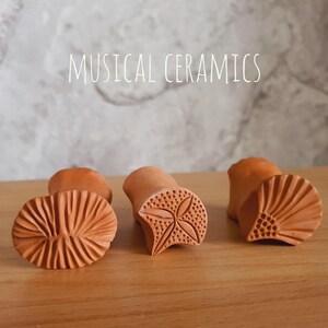 May include: Three terracotta-coloured ceramic stamps with distinct carved patterns on their bases. The left stamp features a radial line design, the centre stamp has a four-petal floral motif, and the right stamp shows a fan-like pattern. Text reads MUSICAL CERAMICS.